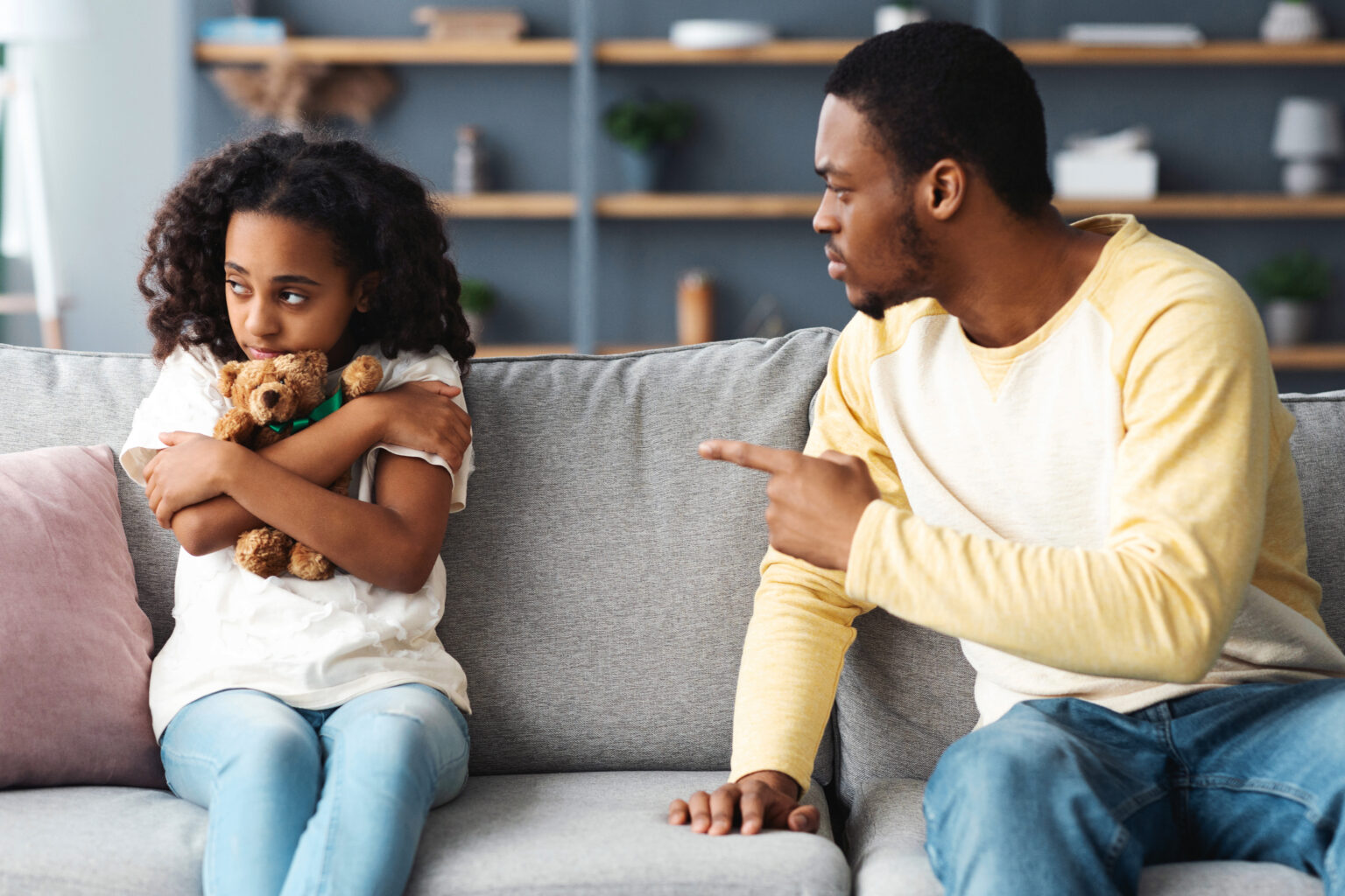 Agressive african american young father yelling at his scared little daughter hugging her teddy bear and looking aside, sitting on sofa together, home interior. Abuse in family concept