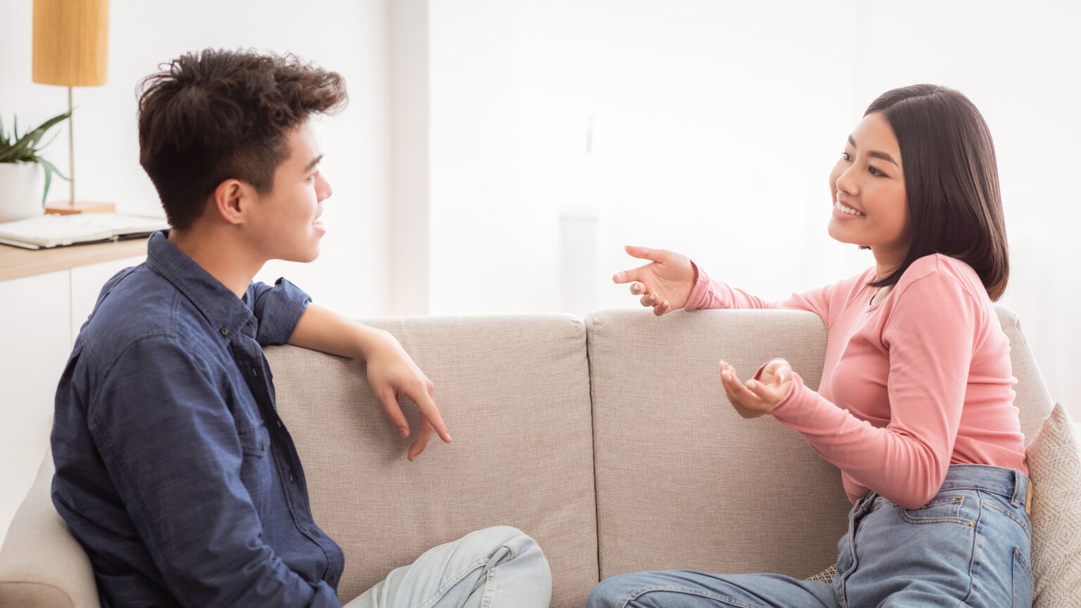 Cheerful Korean Couple Flirting And Talking Enjoying Conversation Sitting On Sofa At Home. Weekend Together, Stay-At-Home Date, Romantic Relationship And Flirt Communication Concept
