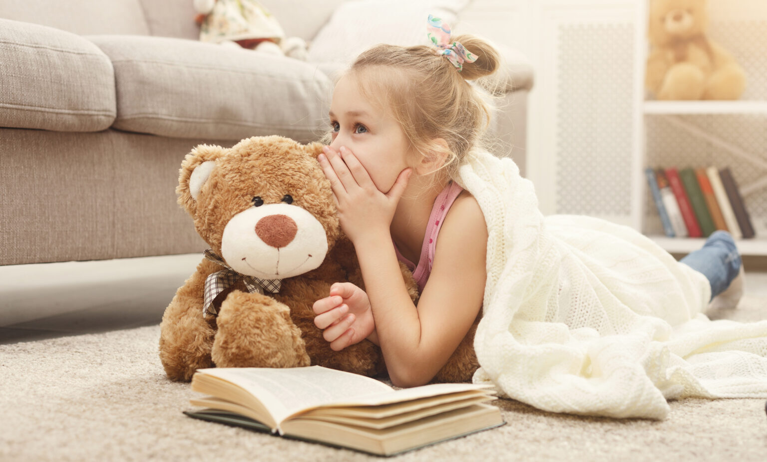 Cute happy little casual girl embracing teddy bear, reading book and sharing secrets with her favorite toy friend. Pretty kid at home, lying on the floor near sofa