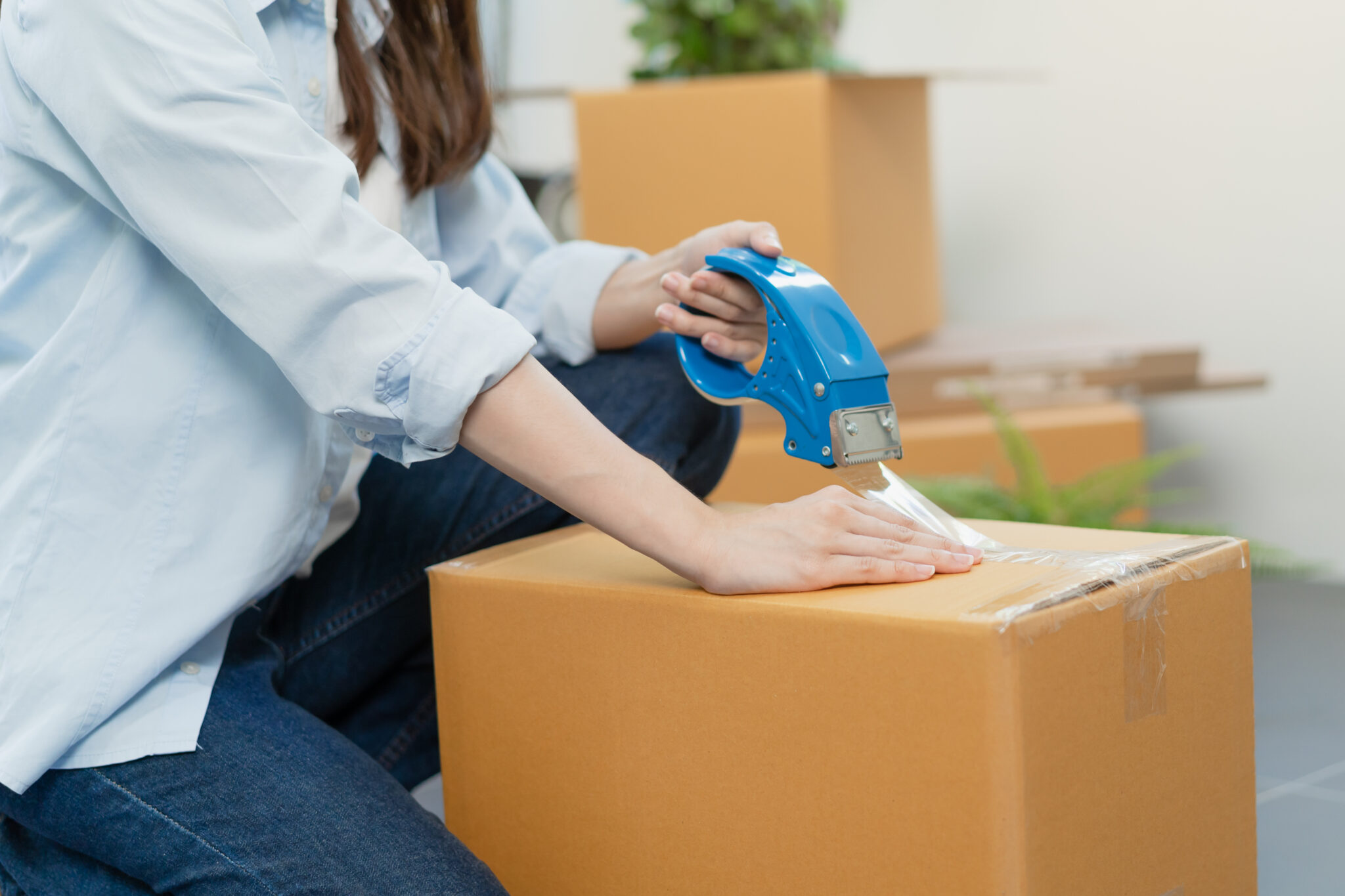 Focus hand of asian woman, female holding packing, unpacking box by tape machine, sealing cardboard indoors. Business owner in warehouse, Moving or preparing in new house, apartment or relocation.