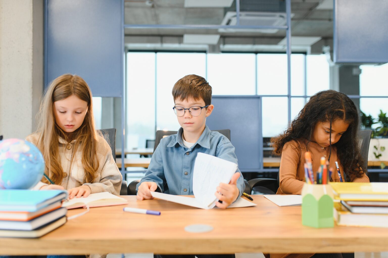 Focused multiracial students kids writing down data into notebook while sitting at table near classmates during test at school